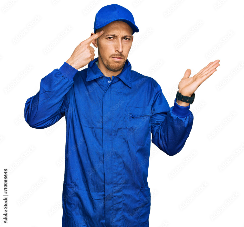 Bald man with beard wearing builder jumpsuit uniform confused and annoyed with open palm showing copy space and pointing finger to forehead. think about it.