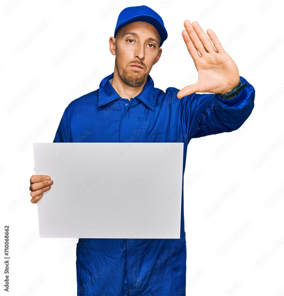 Bald man with beard wearing builder jumpsuit uniform holding empty banner with open hand doing stop sign with serious and confident expression, defense gesture Bald man with beard wearing builder jumpsuit uniform holding empty banner with open hand doing stop sign with serious and confident expression, defense gesture