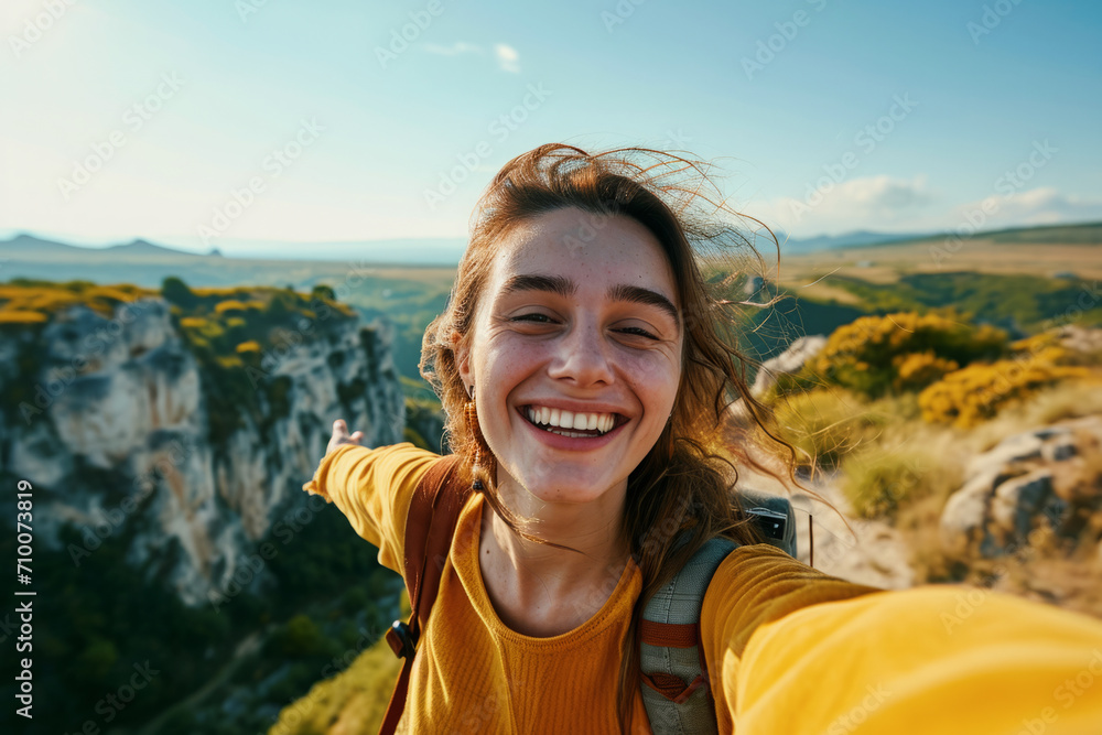 Female traveler with backpack taking selfie against beautiful mountain landscape. Portrait of happy woman hiking in mountains. Travel blogger takes down content