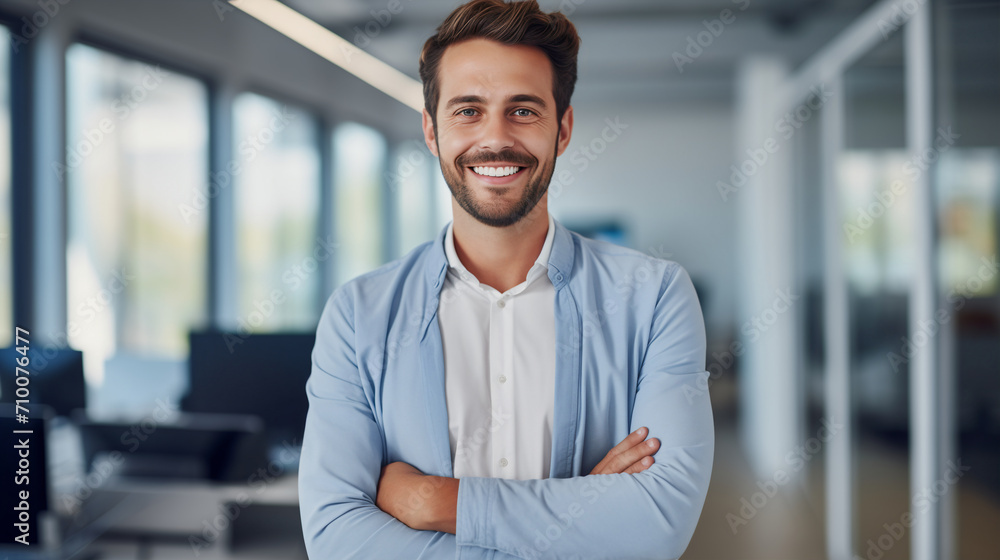 Obraz premium A male freelancer, office employee, man with a captivating smile in smart blue shirt, with his arms crossed in a bright office environment, exudes a blend of professionalism and warmth