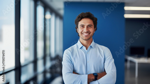 Cheerful man in a business casual blue shirt, arms confidently folded, stands in a contemporary office space, image suitable for themes of innovation, teamwork, and approachable leadership