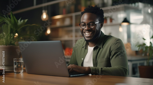 An enthusiastic black young man in a casual olive green shirt is captured laughing while using his laptop, likely reflecting a comfortable and modern co-working space