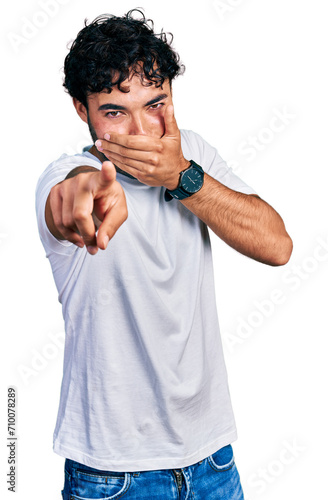 Hispanic young man with beard wearing casual white t shirt laughing at you, pointing finger to the camera with hand over mouth, shame expression
