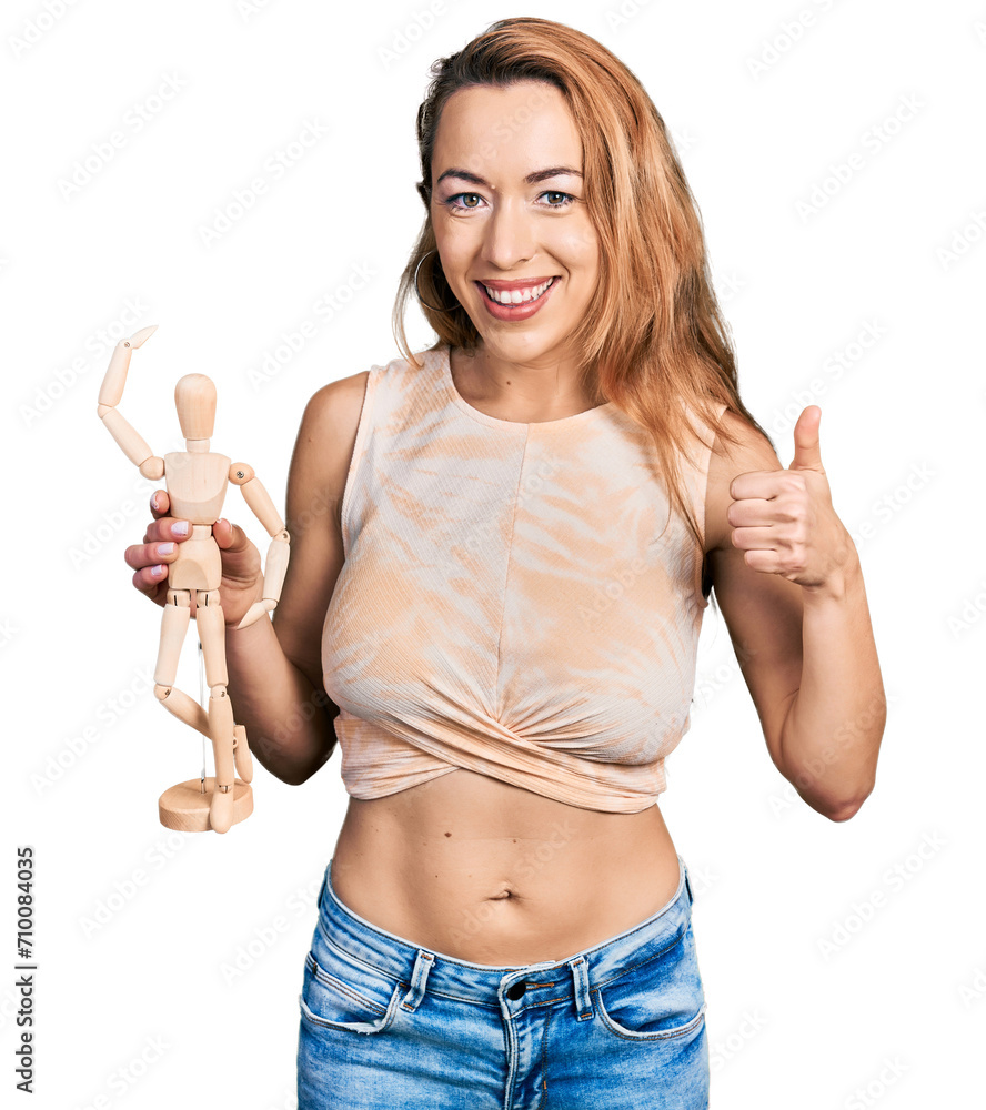 Young caucasian woman holding small wooden manikin smiling happy and positive, thumb up doing excellent and approval sign Young caucasian woman holding small wooden manikin smiling happy and positive, thumb up doing excellent and approval sign