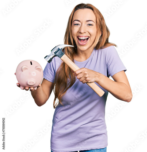 Young caucasian woman holding piggy bank and hammer smiling and laughing hard out loud because funny crazy joke.