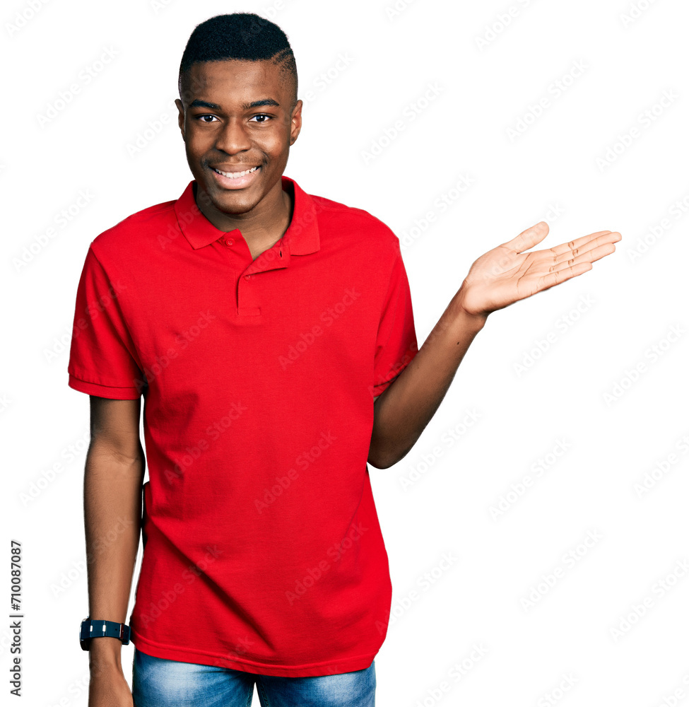 Young african american man wearing casual red t shirt smiling cheerful presenting and pointing with palm of hand looking at the camera. Young african american man wearing casual red t shirt smiling cheerful presenting and pointing with palm of hand looking at the camera.