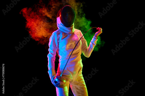 Portrait of female fencer, athlete in white uniform posing with sword over black background in neon light with colorful smoke. Concept of professional sport, competition, championship, hobby © master1305