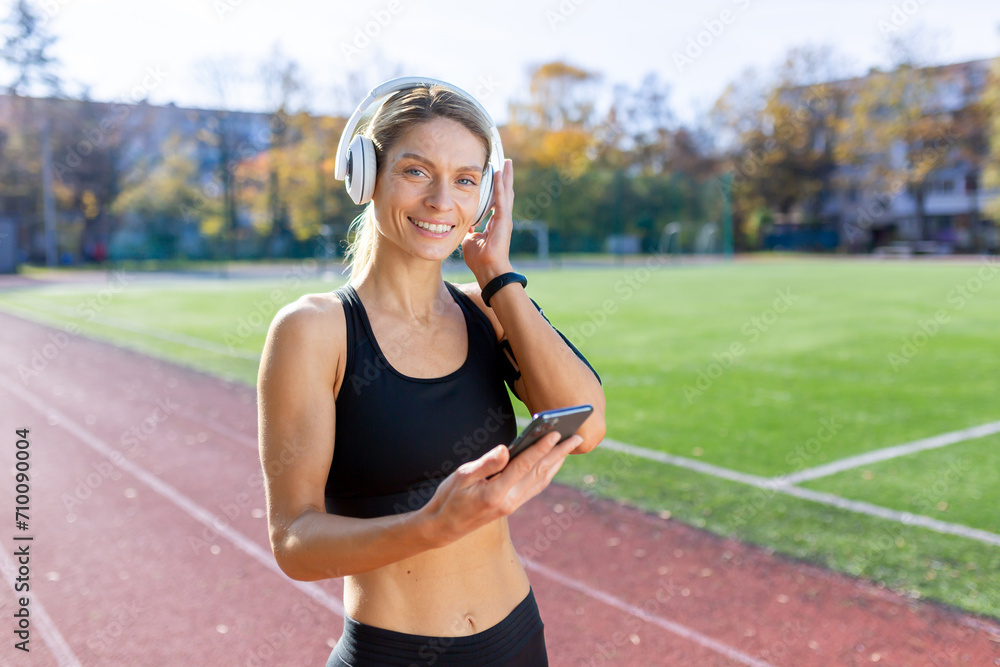 Smiling athletic woman with headphones choosing music on her smartphone at a sunny track field. Concept of fitness and technology. Smiling athletic woman with headphones choosing music on her smartphone at a sunny track field. Concept of fitness and technology.