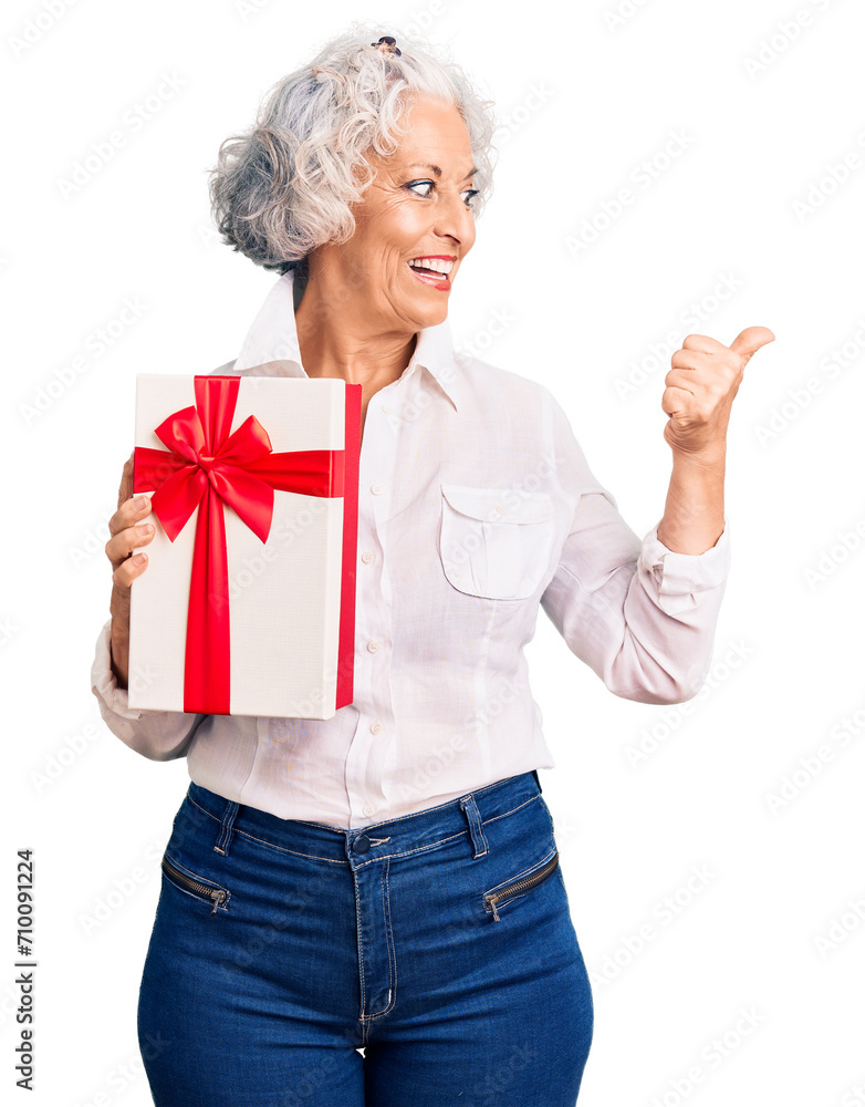 Senior grey-haired woman holding gift pointing thumb up to the side smiling happy with open mouth Senior grey-haired woman holding gift pointing thumb up to the side smiling happy with open mouth