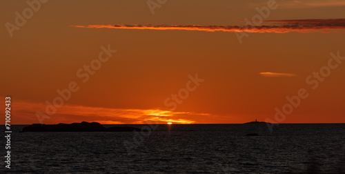 The sun setting in the ocean with small islands and a lighthouse in the horizon.