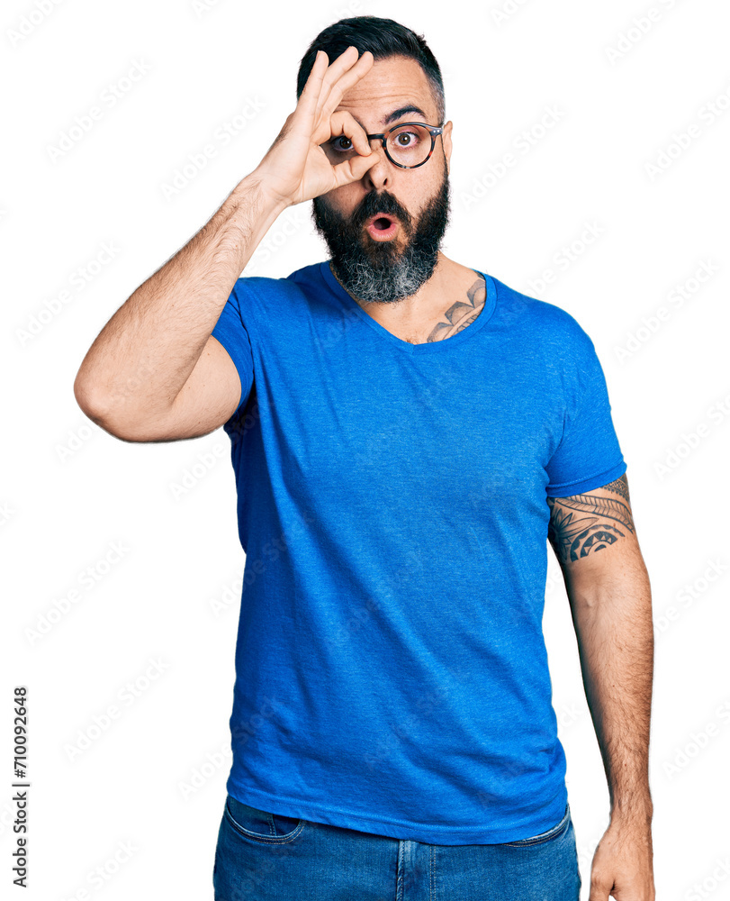 Hispanic man with beard wearing casual t shirt and glasses doing ok gesture shocked with surprised face, eye looking through fingers. unbelieving expression. Hispanic man with beard wearing casual t shirt and glasses doing ok gesture shocked with surprised face, eye looking through fingers. unbelieving expression.