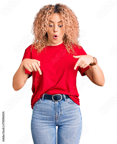 Young blonde woman with curly hair wearing casual red tshirt pointing down with fingers showing advertisement, surprised face and open mouth