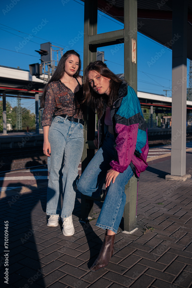 Two beautiful girls dressed in 90s style posing on the platform of the train station on a summer day. Stylish photo of girls dressed in the fashion of the 90s.