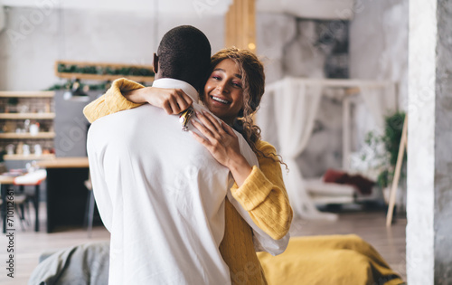 Radiant woman in a yellow sweater embraces man from behind, her face alight with happiness, holding keys to their new dream home, symbolizing a new chapter. Couple new home purchase in dreamy loft