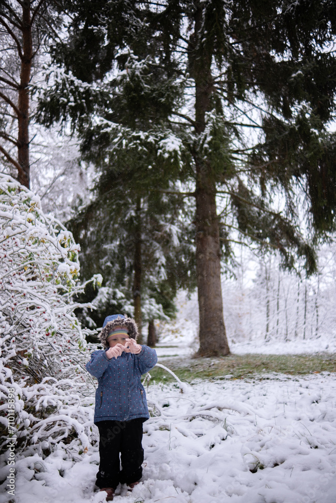 Little beautiful girl in winter clothes standing alone in the middle of a snowy forest. Girl in jacket with fur hood posing in winter forest. Shrubs and trees are covered with snow Little beautiful girl in winter clothes standing alone in the middle of a snowy forest. Girl in jacket with fur hood posing in winter forest. Shrubs and trees are covered with snow