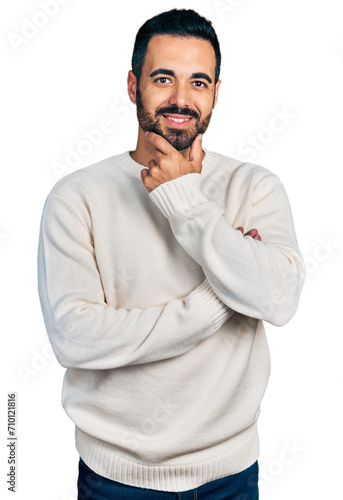 Young hispanic man with beard wearing casual white sweater looking confident at the camera smiling with crossed arms and hand raised on chin. thinking positive.