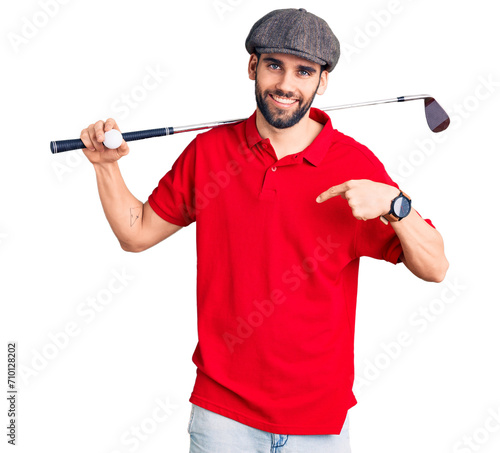 Young handsome man with beard playing golf holding club and ball pointing finger to one self smiling happy and proud