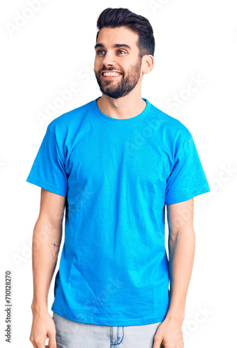 Young handsome man with beard wearing casual t-shirt looking away to side with smile on face, natural expression. laughing confident.