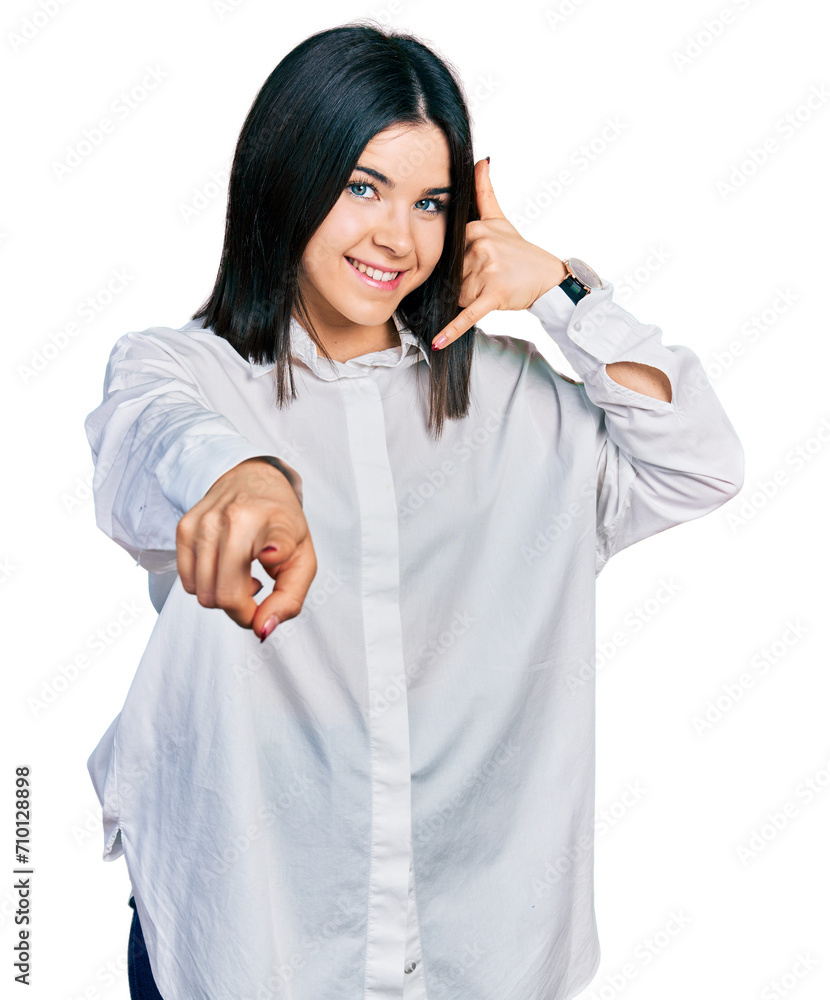 Young brunette woman with blue eyes wearing oversize white shirt smiling doing talking on the telephone gesture and pointing to you. call me.