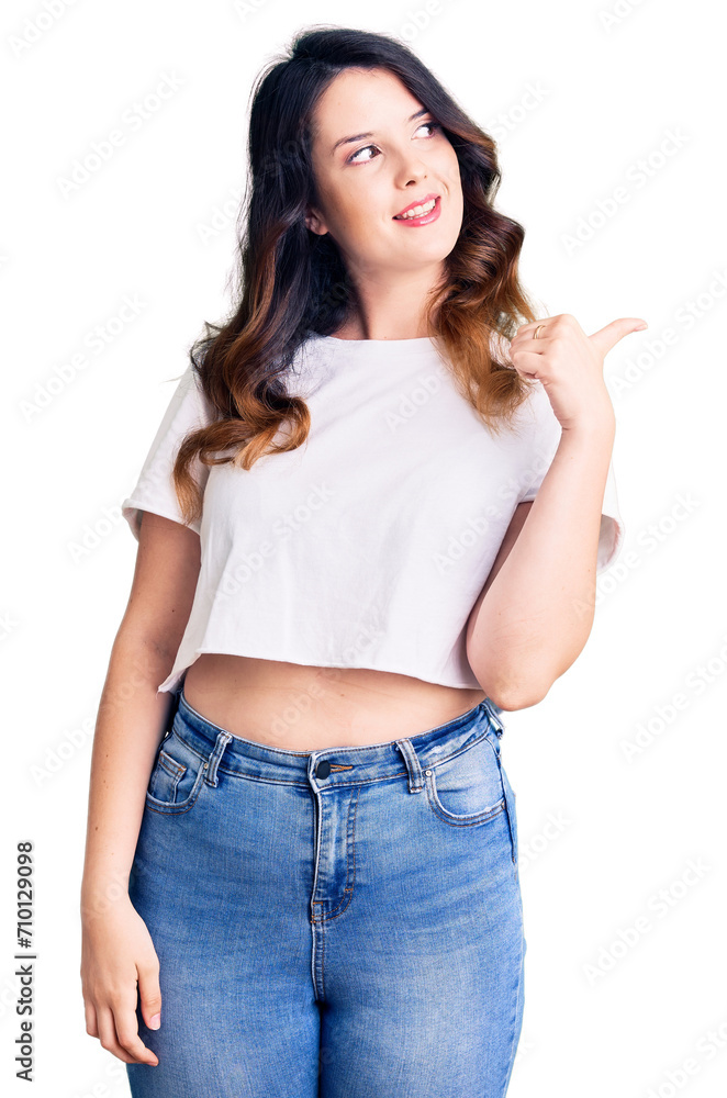 Beautiful young brunette woman wearing casual clothes smiling with happy face looking and pointing to the side with thumb up. Beautiful young brunette woman wearing casual clothes smiling with happy face looking and pointing to the side with thumb up.
