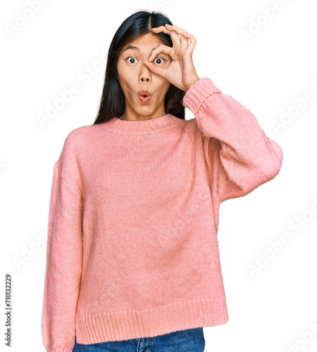 Beautiful young asian woman wearing casual winter sweater doing ok gesture shocked with surprised face, eye looking through fingers. unbelieving expression.
