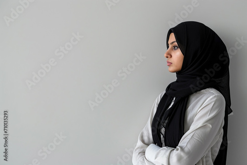 A Muslim girl wearing an Islamic hijab and formal clothes stands against a white wall