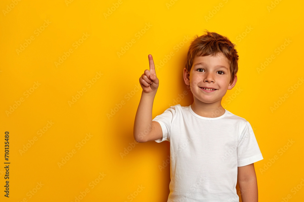 A young boy of Caucasian appearance stands against a yellow wall, pointing and pointing up with his number two finger, smiling a confident and happy smile A young boy of Caucasian appearance stands against a yellow wall, pointing and pointing up with his number two finger, smiling a confident and happy smile