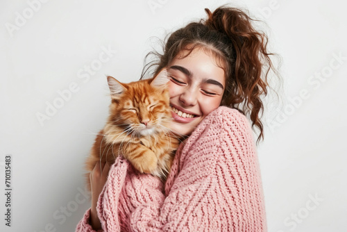 A young beautiful smiling Arab woman in a pink knitted sweater stands on a white background holding and hugging her orange cat. Concept of friendship