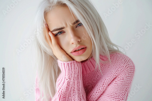 A beautiful blonde girl wearing a pink sweater on a white background is filmed indoors, holding her cheek with her hand, suffering from a severe toothache. © v.senkiv