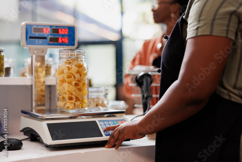 Detailed shot of seller utilizing digital scale to determine weight of container filled with organic produce. Closeup of vendor weighing jar of fresh pasta, with client patiently waiting at counter. photo