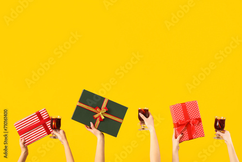 Female hands with glasses of warm mulled wine and gift boxes on yellow background