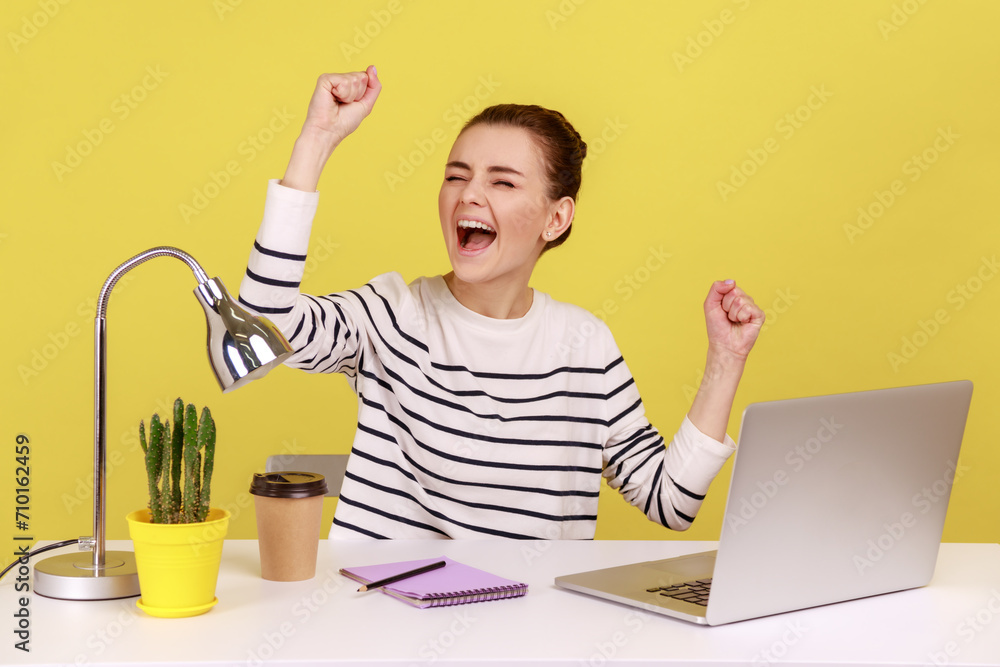 Happy excited young woman office manager raising hands and screaming for joy, celebrating successful business online, sitting at workplace. Indoor studio studio shot isolated on yellow background. Happy excited young woman office manager raising hands and screaming for joy, celebrating successful business online, sitting at workplace. Indoor studio studio shot isolated on yellow background.
