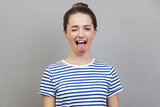Portrait of disobedient crazy funny woman wearing striped T-shirt showing tongue out and closed her eyes displeased, behaving unruly naughty. Indoor studio shot isolated on gray background.