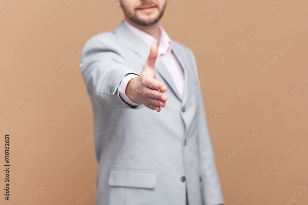 Portrait of unknown man stretching hand for handshaking, accepting job offer, partnership, beginning or end of meeting, wearing gray jacket. Indoor studio shot isolated on brown background. Portrait of unknown man stretching hand for handshaking, accepting job offer, partnership, beginning or end of meeting, wearing gray jacket. Indoor studio shot isolated on brown background.