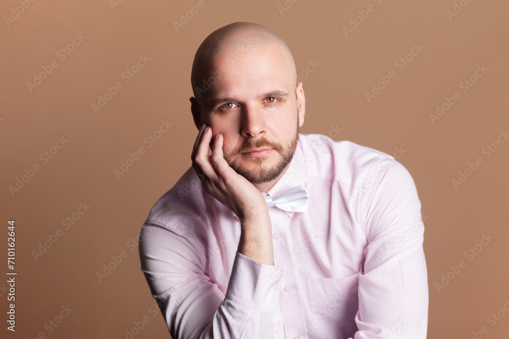 Portrait of calm serious attractive bald bearded man sitting holding his chin, looking at camera, wearing light pink shirt and bow tie. Indoor studio shot isolated on brown background. Portrait of calm serious attractive bald bearded man sitting holding his chin, looking at camera, wearing light pink shirt and bow tie. Indoor studio shot isolated on brown background.