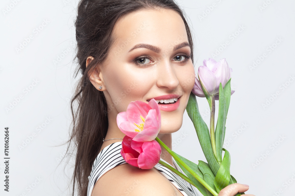 Side view portrait of smiling beautiful brunette woman holding bouquet of tulips, looking at camera, smiling with happiness, wearing striped dress. Indoor studio shot isolated on gray background. Side view portrait of smiling beautiful brunette woman holding bouquet of tulips, looking at camera, smiling with happiness, wearing striped dress. Indoor studio shot isolated on gray background.