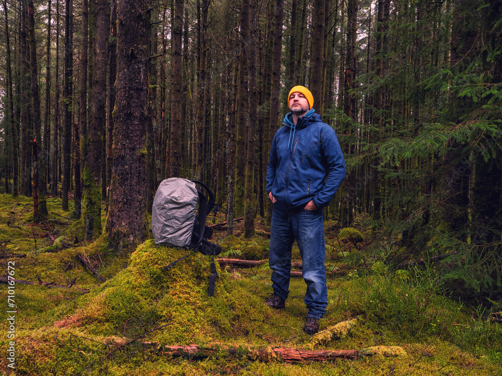 Naklejka premium Male tourist with backpack covered with rain cover in a dense forest. Man in blue jeans and jacket and yellow warm hat. Travel and explore nature concept. Outdoor trip theme.