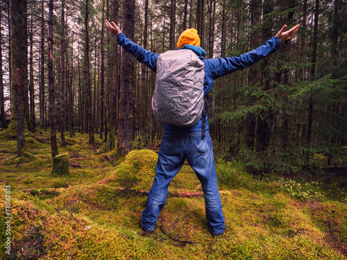Male tourist with backpack covered with rain cover in a dense forest. Man in blue jeans and jacket and yellow warm hat. Travel and explore nature concept. Outdoor trip theme. Hands up gesture.