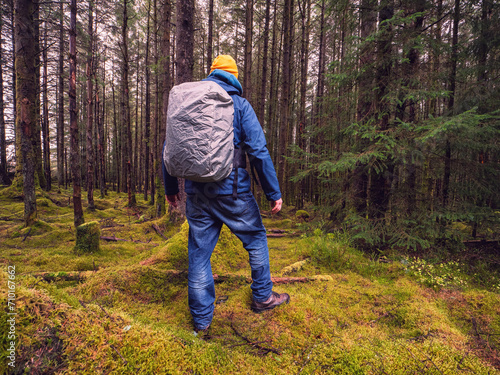 Male tourist with backpack covered with rain cover in a dense forest. Man in blue jeans and jacket and yellow warm hat. Travel and explore nature concept. Outdoor trip theme.