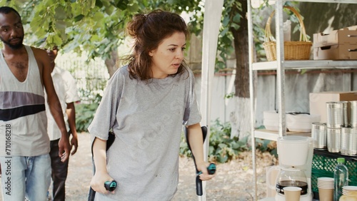 Poor, hungry Caucasian lady on crutches approaches an outdoor food bank to receive free food assisted by black woman. Volunteers serving meals and nourishments to underprivileged and homeless people.
