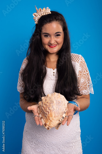 Beautiful young woman in white dress with shell tiara on her head holding a sea shell and looking at the camera.