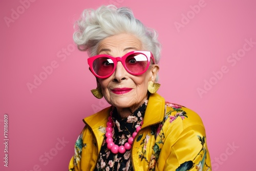 Fashionable senior woman in bright clothes and sunglasses posing over pink background. © Iigo
