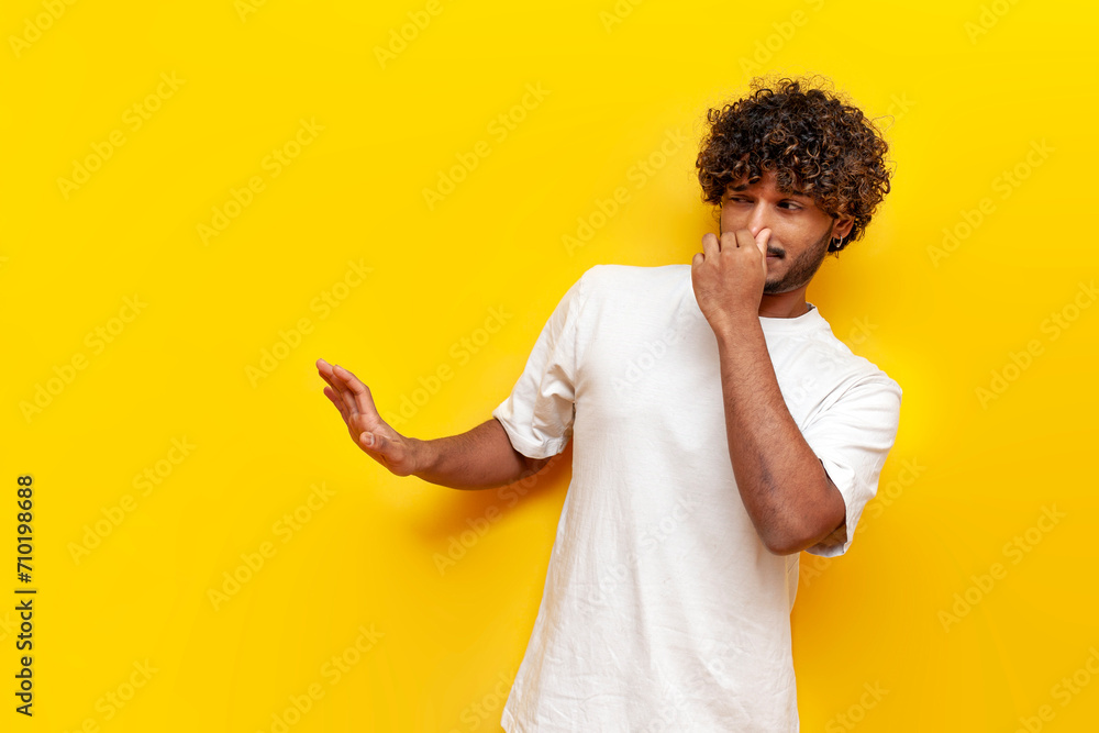 young curly indian man showing disgust and covering his nose with hand on yellow isolated background, guy refuses bad smell and rejects stench young curly indian man showing disgust and covering his nose with hand on yellow isolated background, guy refuses bad smell and rejects stench