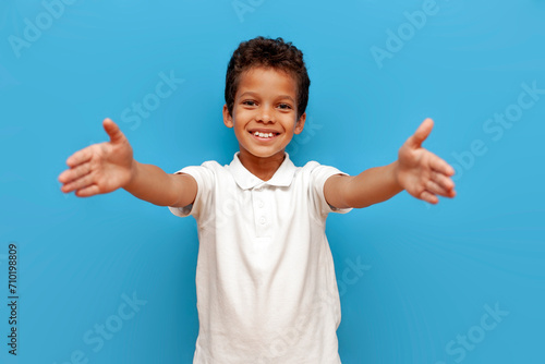 African American boy in white polo wants to hug and pulls his hands forward on blue isolated background, 10 year old child holds empty hands in front of him photo