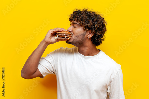 young hungry indian guy eating delicious cheeseburger on yellow isolated background, curly man eating fast food and biting burger
