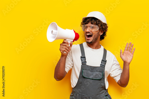 young indian male builder in a hard hat and overalls announces information into a loudspeaker on a yellow isolated background, a foreman in a uniform and safety glasses shouts into a megaphone