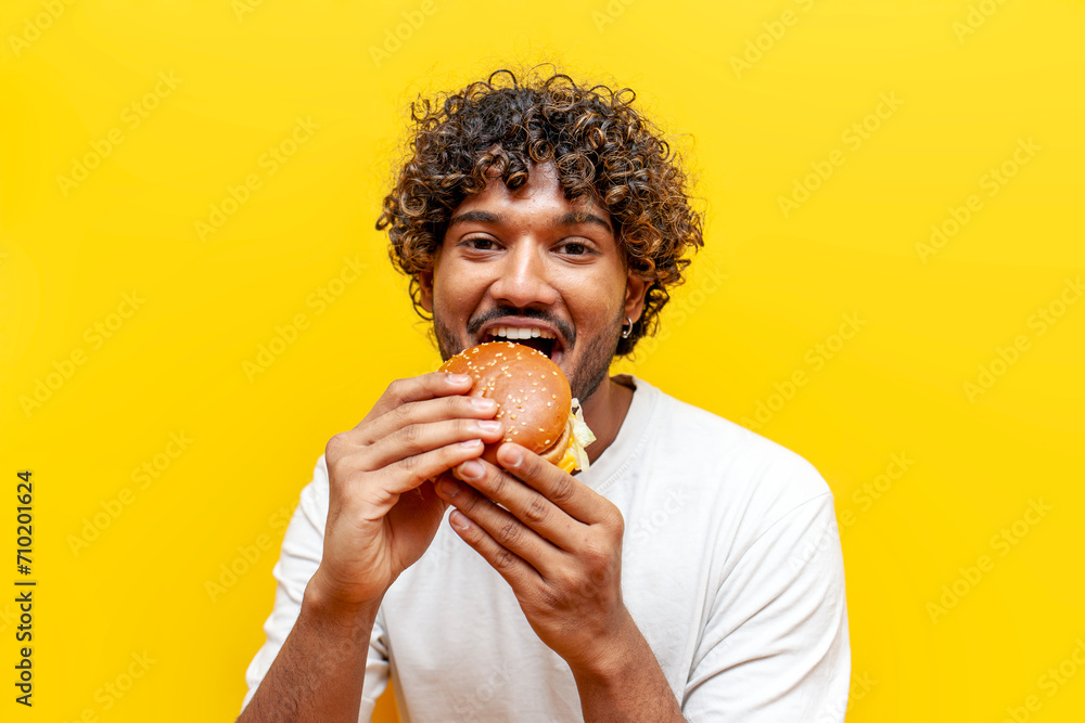 young hungry indian guy eating delicious cheeseburger on yellow isolated background, curly man eating fast food and biting burger young hungry indian guy eating delicious cheeseburger on yellow isolated background, curly man eating fast food and biting burger