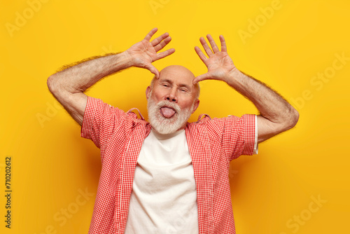 cheerful old bald grandfather in summer shirt is fooling around and shows his tongue and horns on yellow isolated background, crazy elderly pensioner is grimacing and joking