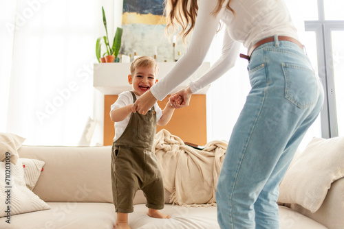 young mother and her little son dance and jump on the sofa at home  2-year-old boy screams and rejoices with his parent  woman has fun with her child