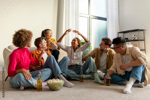 multiracial group of young people sitting at home with beer and popcorn and playing charades and having fun with friends, students of different ethnicities at home playing games and celebrating photo